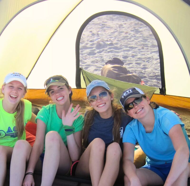 The image shows four young women inside a tent, likely camping on a beach. They are all smiling and appear to be enjoying themselves. The woman on the left is wearing a yellow shirt and a white cap, while the others are wearing blue or green shirts and caps or sunglasses. The background shows a sandy beach and possibly the ocean.